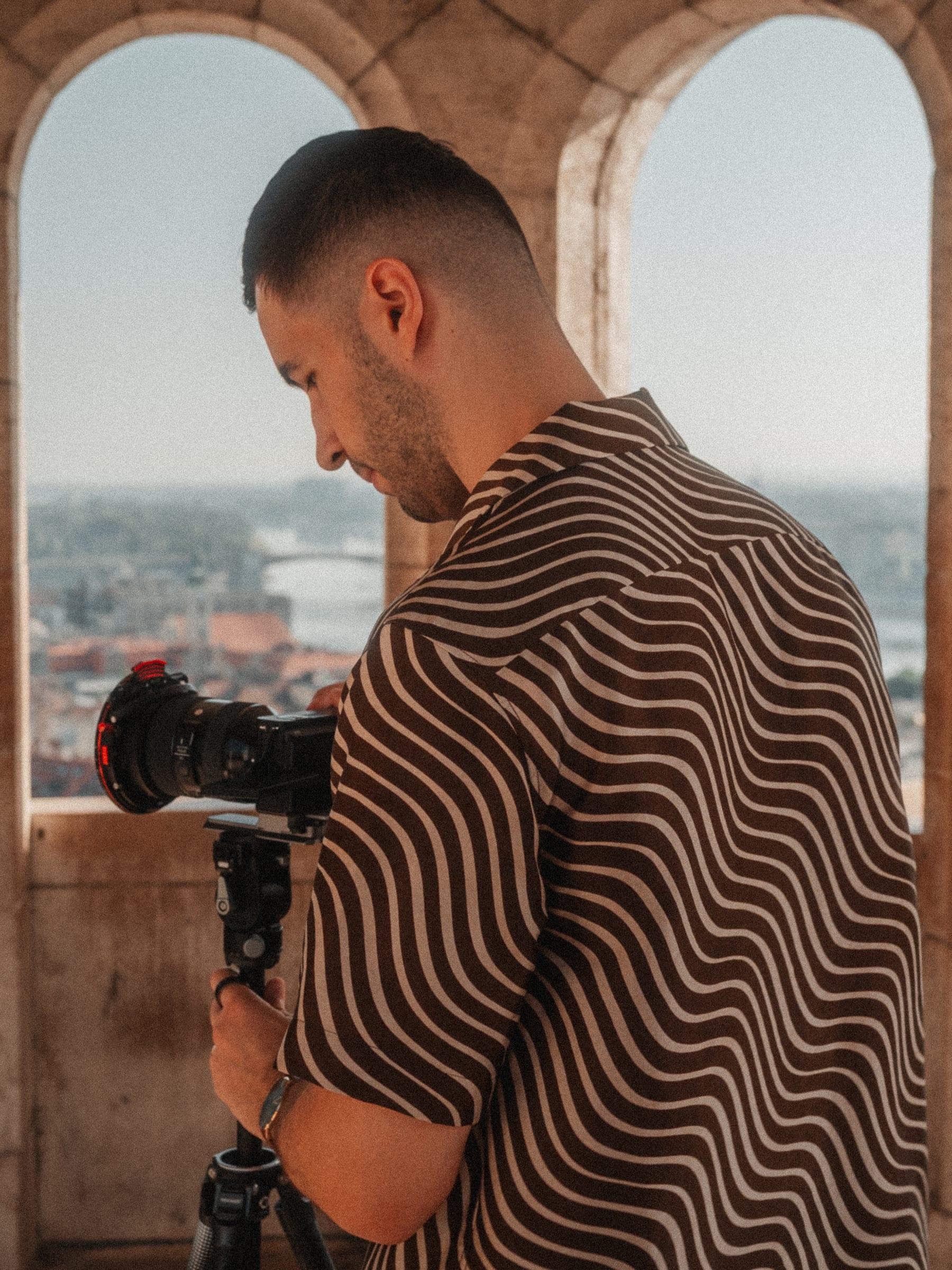 A camera operator at a tripod inside a stone arch, the city behind