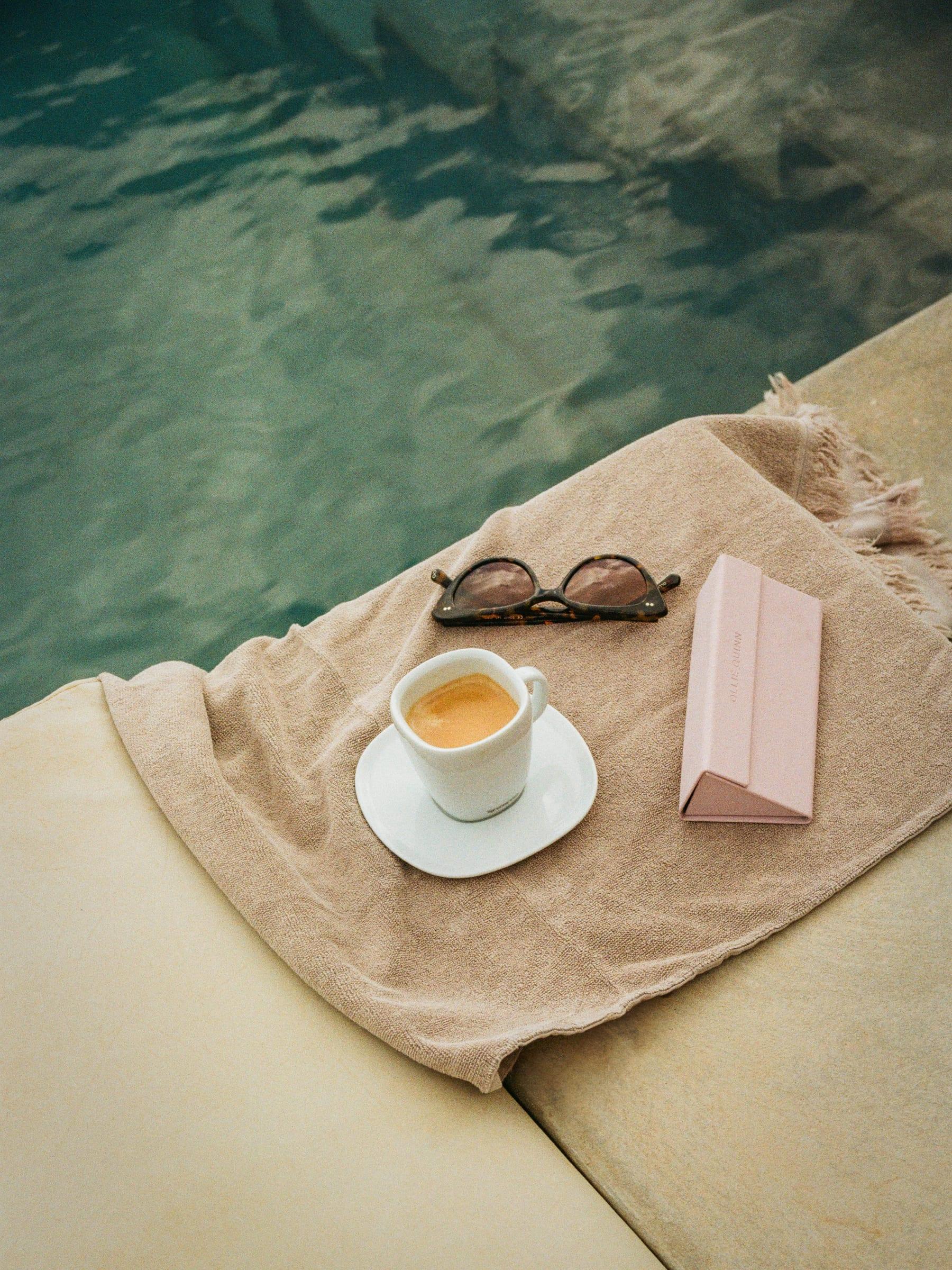 An espresso and a pair of sunglasses left on the towel at the water’s edge